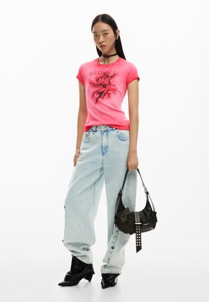 Vrouw draagt een roze T-shirt met print, lichtblauwe wijde jeans, zwarte puntige laarzen en houdt een zwarte bloemenhandtas met zilveren accenten vast.