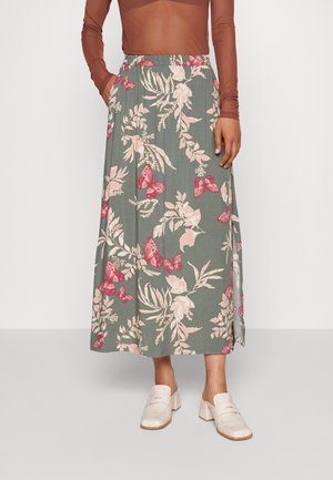 Lange Blumenrock aus grünem Stoff, mit rosa Schmetterlingen und Blättern. Elastischer Bund, seitliche Taschen, kombiniert mit hellen Mules.