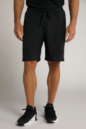 Mand iført sorte træningsshorts med snøre og sorte løbesko står på en ensfarvet baggrund.