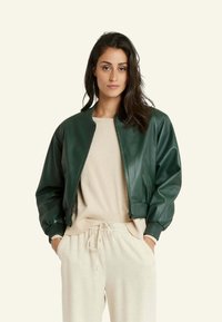 Veste bomber en cuir vert avec des poignets et un ourlet côtelés, fermeture éclair, portée sur un pull en tricot beige et un pantalon clair. Texture lisse.