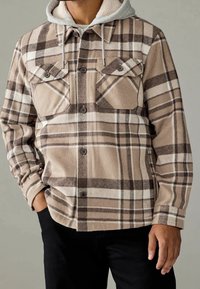 Beżowa koszula flanelowa w kratę z brązowymi i białymi paskami, z dwoma kieszeniami na piersi, zapięciem na guziki i jasnoszarym wykończeniem w kapturze.
