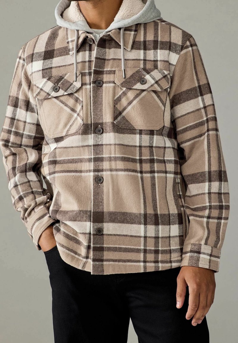 Beżowa koszula flanelowa w kratę z brązowymi i białymi paskami, z dwoma kieszeniami na piersi, zapięciem na guziki i jasnoszarym wykończeniem w kapturze.