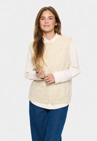 Beige stickad väst med rund hals, bärs över en vit skjorta med knappar. Slät textur, avslappnad passform, lager på lager över blå denimbyxor.