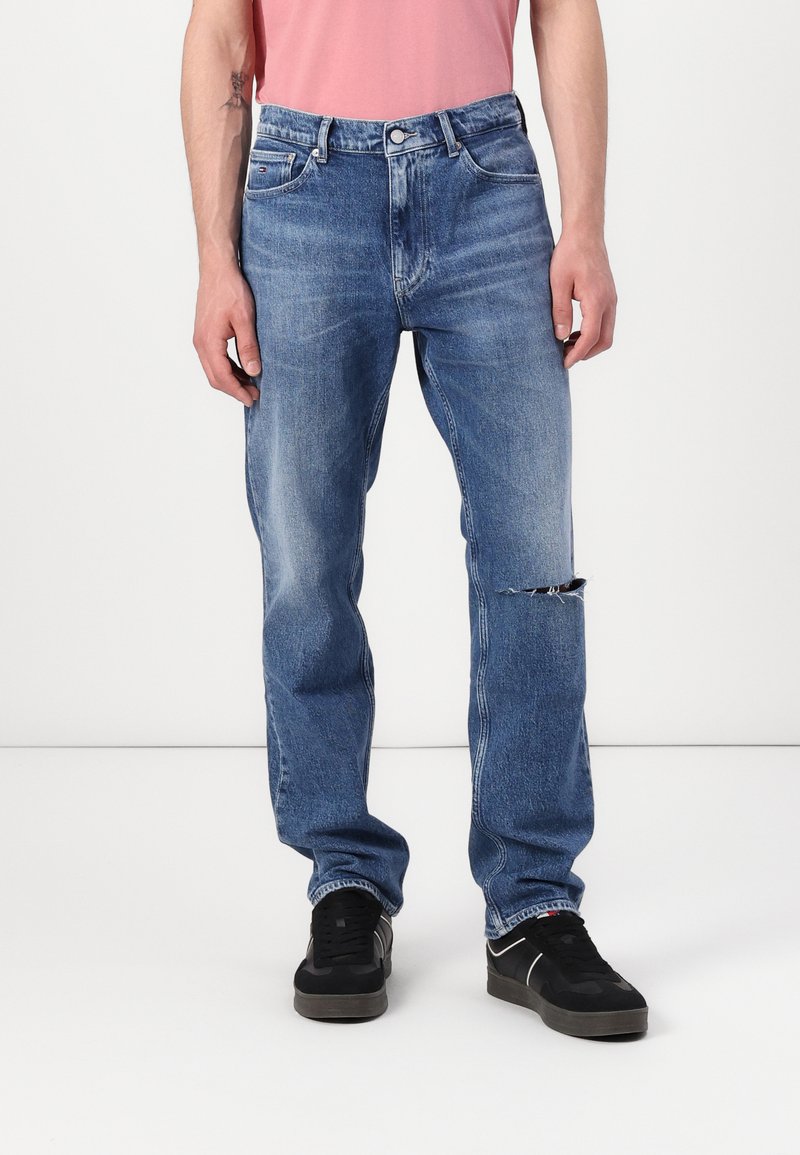 Vaqueros de mezclilla azul claro con corte recto, cintura de altura media y un desgastado en la rodilla izquierda. Combinados con zapatillas negras.