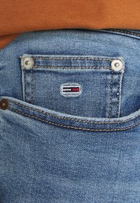 Vaqueros de mezclilla de color azul claro, con un pequeño bolsillo, herrajes plateados y una etiqueta con el logotipo que presenta rayas rojas y negras. Tejido texturizado.