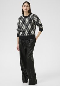 Cardigan argyle noir et blanc avec un col rond, fermeture à boutons et manches côtelées, associé à un pantalon large noir brillant.