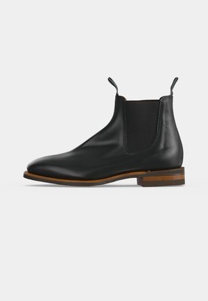 Botte Chelsea en cuir noir avec panneaux latéraux élastiques, languettes de tirage à la cheville et petit talon superposé marron, présentée de profil sur fond blanc.