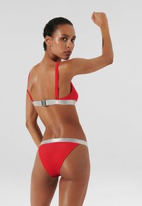 Bikini rosso con dettagli argentati. Il top ha spalline regolabili e chiusura sul retro. La parte inferiore presenta un design a vita bassa con una cintura coordinata.
