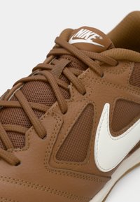 Smijeđa tenisica s teksturiranim kožnim gornjištem, mrežastim detaljima i bijelim Nike swoosh logom. Ima smeđe vezice i podstavljeni ovratnik.