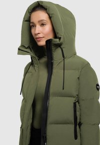 Donna che indossa una giacca imbottita verde oliva con un ampio cappuccio e dettagli in zip neri, che guarda leggermente di lato su uno sfondo grigio chiaro.