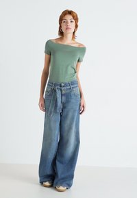 Grønn av-shoulder topp kombinert med oversized lys blå denimjeans, med knyttet beltedetalj. Modellen står foran en nøytral bakgrunn.