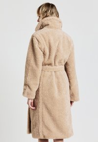 Accappatoio beige in morbido tessuto, con colletto alto, maniche lunghe e cintura in vita, caratterizzato da un tessuto texturizzato e peloso e un design di lunghezza intermedia.