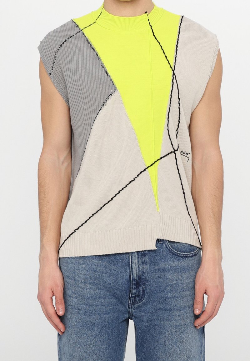 Débardeur en tricot sans manches avec des blocs de couleur abstraits gris, beige et jaune fluo, des lignes noires, porté avec un jean bleu par une personne debout.