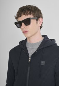 Jonge man met zwarte geometrische zonnebril, een donkere gebreide hoodie met rits en logo patch, en een grijze hoge hals t-shirt, tegen een effen achtergrond.