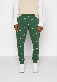 Gröna sweatpants i bomull med en mjuk textur, prydda med spridda vita Nike-logotyper och en tapered design, kombinerade med vita sneakers.