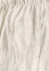 Ljus beige tyg med en rynkig textur, med en ficka med lappar och en liten sydd etikett med "STREET ONE STUDIO" i vitt.