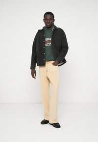 Veste en denim noire sur un hoodie vert avec du texte, pantalon large crème, et chaussures noires. Fond blanc, axé sur les détails des vêtements.