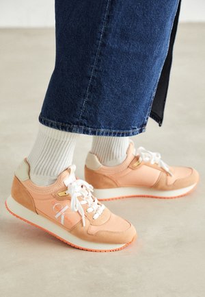 Osoba ubrana w białe skarpetki, niebieskie dżinsy i brzoskwiniowe sneakersy Calvin Klein stojąca na jasnej podłodze.