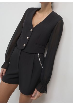 Czarny, dopasowany kombinezon z przezroczystymi, długimi rękawami, perłowymi guzikami z przodu i perłowymi zdobieniami przy prawym kieszeni, noszony przez stojącą osobę.