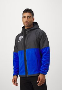 Sort og blå fleecejakke med hætte, med lynlås, to sidelommer og et logo på den øverste venstre side.