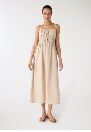 Donna in piedi che indossa un abito midi beige senza maniche con corpetto arricciato e spalline sottili, abbinato a sandali piatti con cinturini.