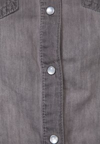 Camicia in denim grigio con cinque bottoni a pressione argentati lungo la parte frontale, caratterizzata da una consistenza morbida e doppie tasche sul petto con dettagli di cucitura sottili.