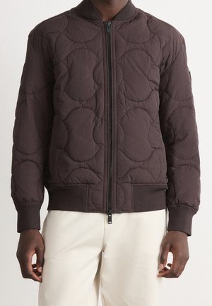 Mann trägt eine dunkelbraune gesteppte Jacke mit Frontreißverschluss und gerippten Bündchen, kombiniert mit hellbeiger Hose.