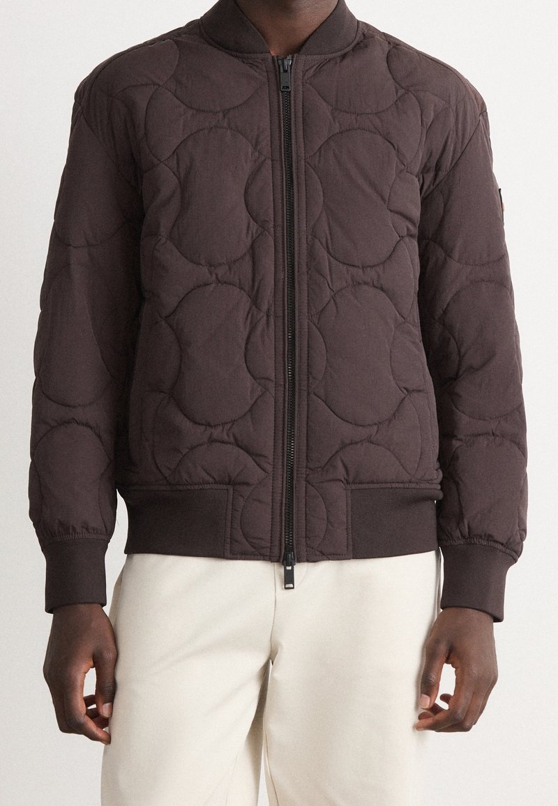 Homme portant une veste matelassée marron foncé avec une fermeture éclair à l'avant et des poignets côtelés, associée à un pantalon beige clair.