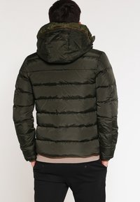 Oliven grøn pufferjakke med quiltet design, hætte og ribbet kant. Har en camouflageforing og glat tekstur.