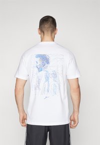 Hvit bomulls t-skjorte med et blått grafisk design av en stilisert figur og geometriske linjer på baksiden. Korte ermer og rund hals.