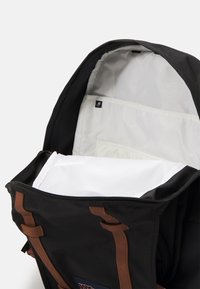 Zaino nero con spallacci in pelle marrone; l'interno presenta scomparti bianchi e un'etichetta; tessuto robusto con chiusura a zip.