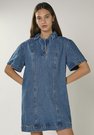 Jeune femme aux longs cheveux foncés portant une robe en jean bleue à manches courtes avec un col et des détails de couture à l'avant.