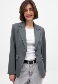 Grå blazer med skræddersyet pasform, enkelt knaplukning, spidse reverser og forlommer. Båret over en hvid top og parret med grå jeans.