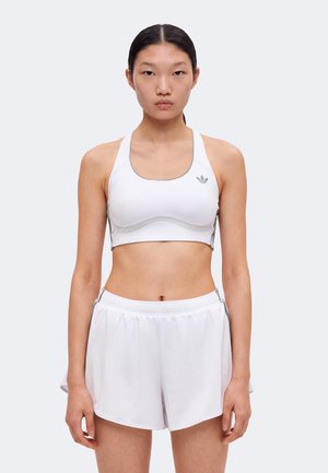 Ung kvinde står iført en hvid sports-bh og matchende atletikshorts mod en ensfarvet lys baggrund.