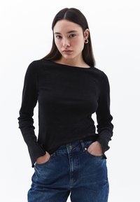 Schwarzes Langarmoberteil mit Boot-Ausschnitt und strukturierter Oberfläche, kombiniert mit hochgeschnittenen blauen Jeans.