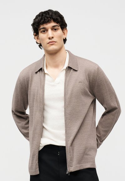 Νέος άνδρας με κοντά σγουρά μαλλιά, ντυμένος με μπεζ ζιπ-αρ καρντιγκάν πάνω από λευκό πουκάμισο και μαύρο παντελόνι, στέκεται με τα χέρια πίσω από την πλάτη.