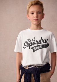 Blond pojke som bär en vit "Real Superdry" t-shirt med japanska tecken, beige byxor och en marinblå tröja knuten runt midjan.