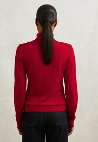 Maglione rosso lavorato a maglia con colletto alto, maniche lunghe e spalline sulle spalle. La vestibilità è aderente, con un orlo e polsini a coste.