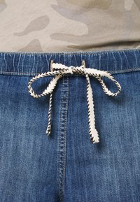 Shorts de mezclilla con un lavado azul, que presentan un cierre de cordón de tela crema con un patrón negro y una textura suave.