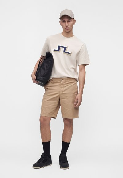 Homme portant une casquette beige, un t-shirt graphique beige, un short kaki, des chaussettes noires, des baskets, tenant un sac à dos noir sur fond blanc.