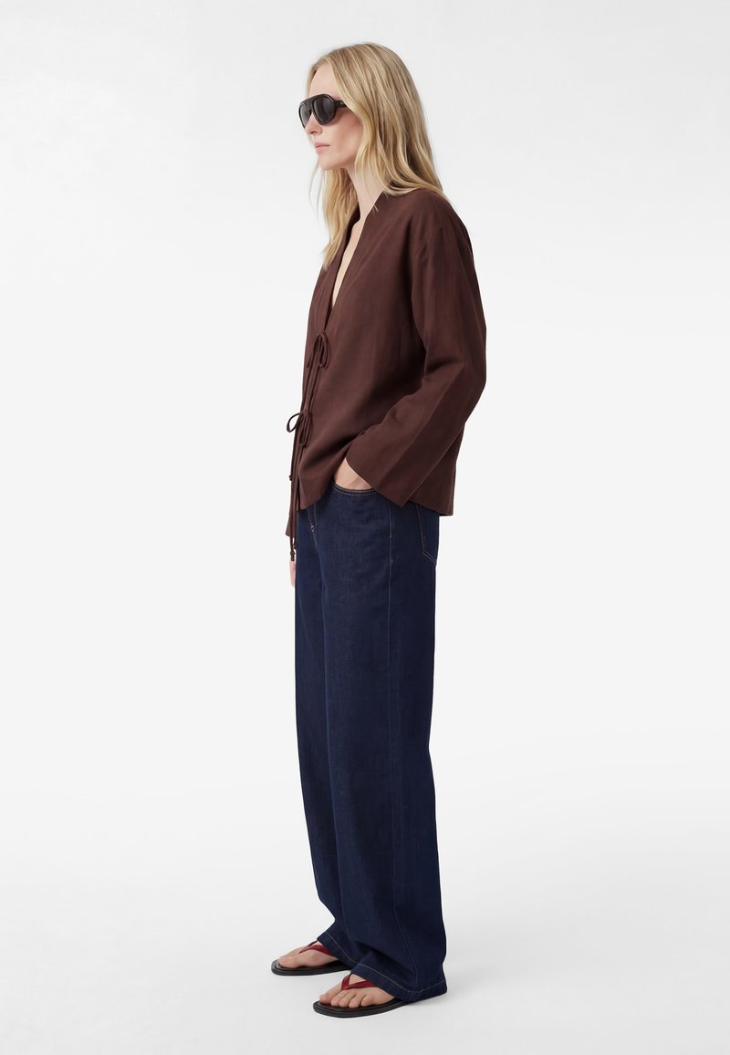 Frau mit dunkler Sonnenbrille, braunem, langärmligem, gebundenem Top, weiten marineblauen Hosen und roten Flip-Flops, steht mit einer Hand in der Tasche.