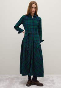 Vestido de manga larga a cuadros en verde y azul marino, con cierre de botones en el frente, cintura entallada y falda acampanada. Estilizado con zapatos marrones.