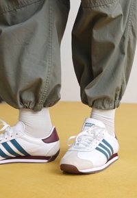 Balti sportiniai batai su mėlyni stogai ir raudonai ruda akcentais; su raišteliais; nubuko ir lygūs paviršiai; dėvimi su žaliais elastiniais kelnais.