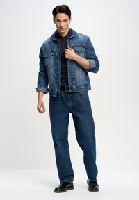 Mėlynas džinsinis švarkas klasikinio kirpimo, uždėtas ant juodo polo marškinėlio. Sukombinuota su šviesiai mėlynomis tiesiomis kelnėmis ir juodais batais.