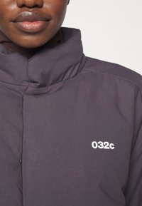 Gros plan d'une personne portant une veste matelassée foncée avec un col montant et un logo blanc "032c" sur la poitrine.
