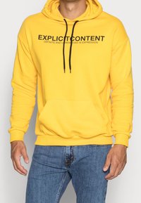 Żółta bluza z kapturem wykonana z mieszanki bawełny, z przednią kieszenią typu kangur oraz czarnymi sznurkami, na piersi z odważnym nadrukiem tekstowym.