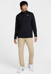 Μαύρο μακρυμάνικο πουλόβερ με φερμουάρ στον λαιμό, με λευκό λογότυπο Nike. Συνδυασμένο με μπεζ παντελόνι και λευκά αθλητικά παπούτσια. Λείος υφής.