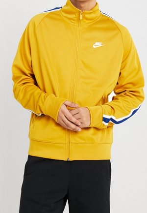 Mand iført en sennepsgul Nike lynlåsjakke med blå og hvide striber på ærmerne, hænderne foldet foran, kombineret med sorte bukser.