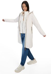 Hellbeige wasserdichte Jacke mit verstellbarem Kapuzen, Frontreißverschluss und zwei Seitentaschen, getragen über einem weißen Fleece-Oberteil und blauen Jeans.