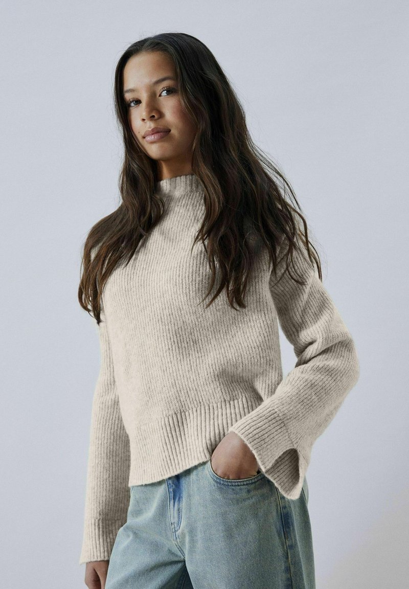 Maglione a coste beige con alto collo e polsini a spacco, abbinato a jeans blu chiaro. Texture morbida, design aderente.